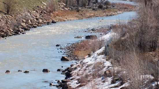 Muddy flowing river with large rocks around the bank pressed against dead brush on a winter day.