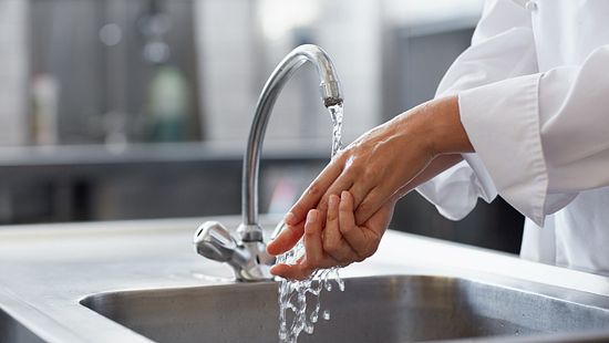 Person in a white long-sleeved garment washing hands under a running faucet in a stainless steel sink, with a blurred kitchen or industrial background.