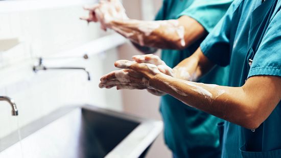 Two healthcare workers in teal scrubs wash their hands with soap at a stainless-steel sink in a clinical setting.