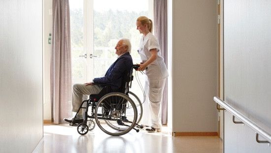 A health worker pushing the wheelchair of an elderly man dressed in a complete suit across a hospital hallway.