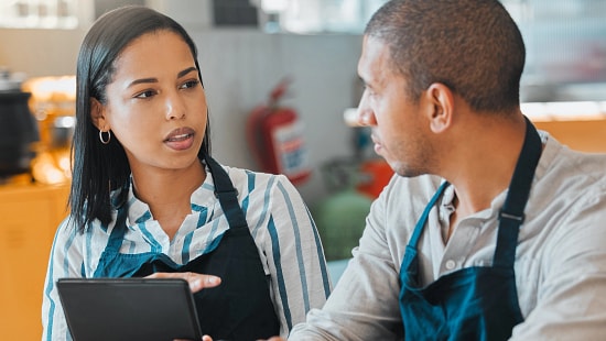 Two quick service restaurant workers talking, one is holding a digital tablet.
